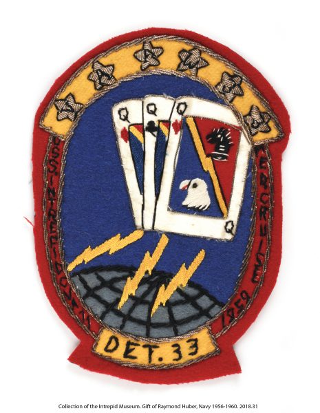 The Squadron VA(AW)-33 Mediterranean Cruise Patch is an oblong patch associated with all-weather attack squadron VA(AW)-33 detachment 11. The patch has a red border with six stars spelling "VAAW 33" above center image and "DET. 33" below. Both phrases are on arched yellow backgrounds. In the center are three queen cards: queen of diamonds, hearts and clubs. The face of the top card has a yellow lightning bolt diagoally down the center. The lower section has a bluew background with egale head and the upper ection has a red background with black knight chess piece (horse's head). Below the cards are three yellow lightning bolts shooting toward a gray globe. The image background is dark blue. "USS Intrepid CVA 11 // Med. Cruise 1959" is spelled vertically on either side of the center image. The borders and stars have metallic embroidery. The patch has a gray backing.