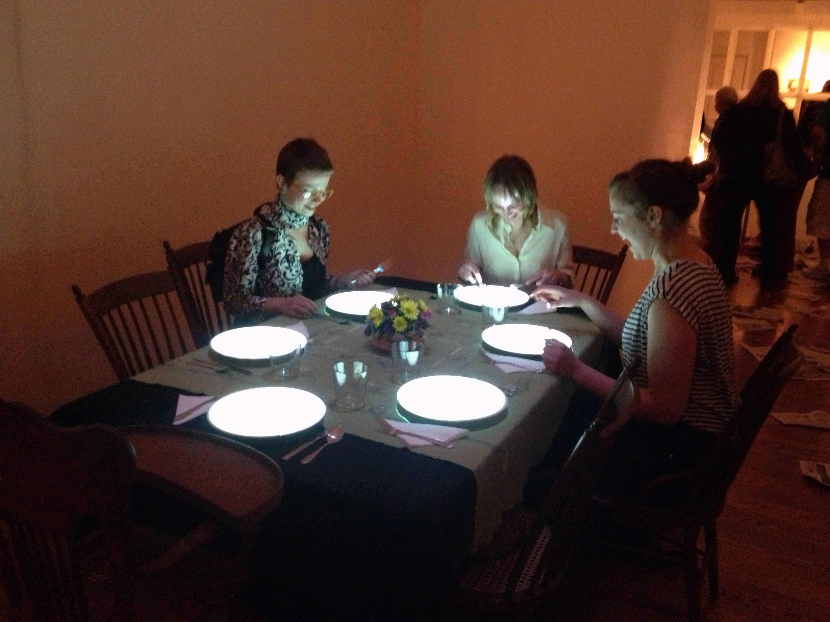 A video installation about hunger and chance - a photo of three people in sitting around a dinning table set for six, with plates glowing with video images, their faces lit from the glow