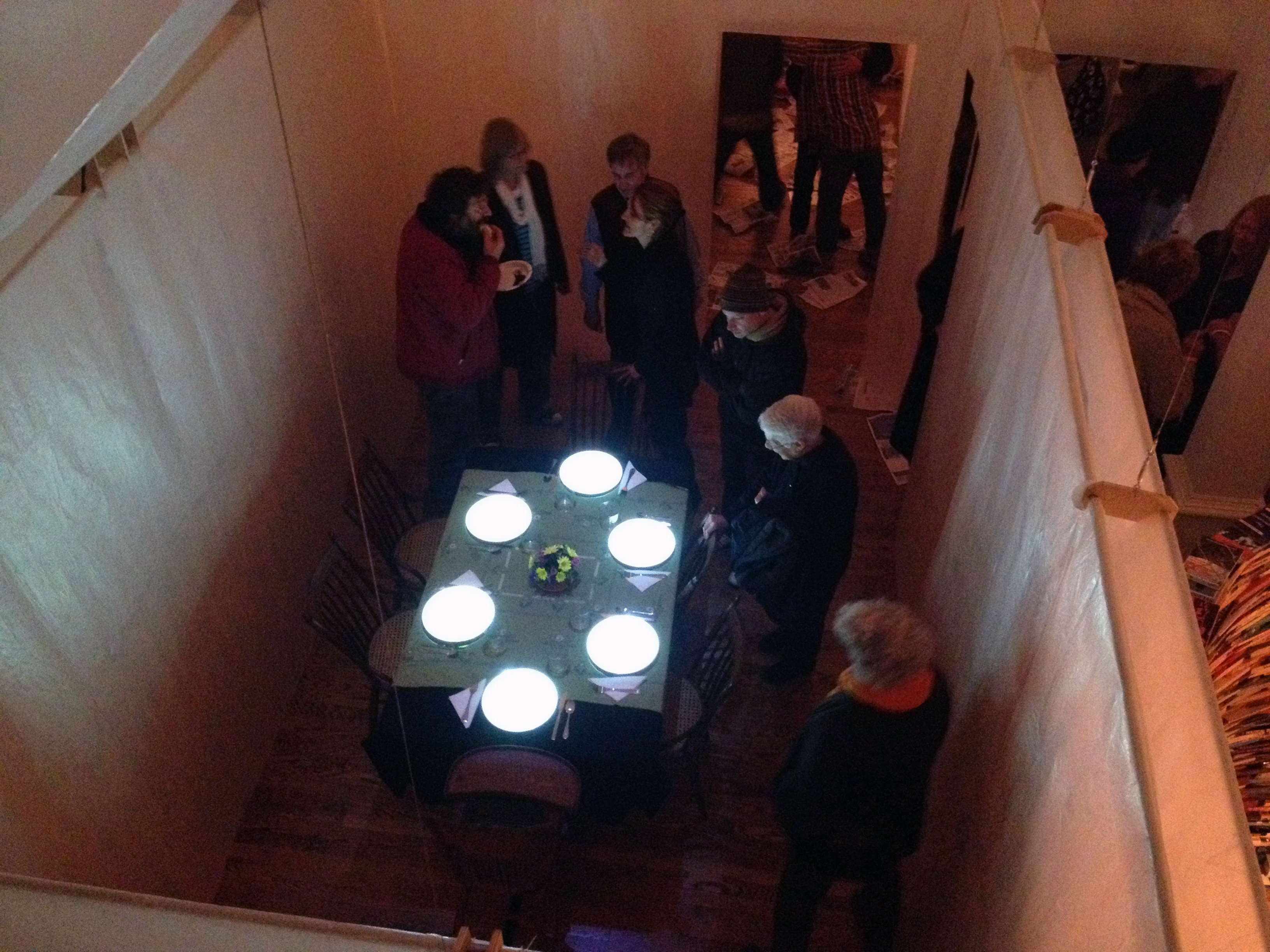 A video installation about hunger and chance - a photo of the installation from above, there is a crowd around a dinning table set with glowing plates