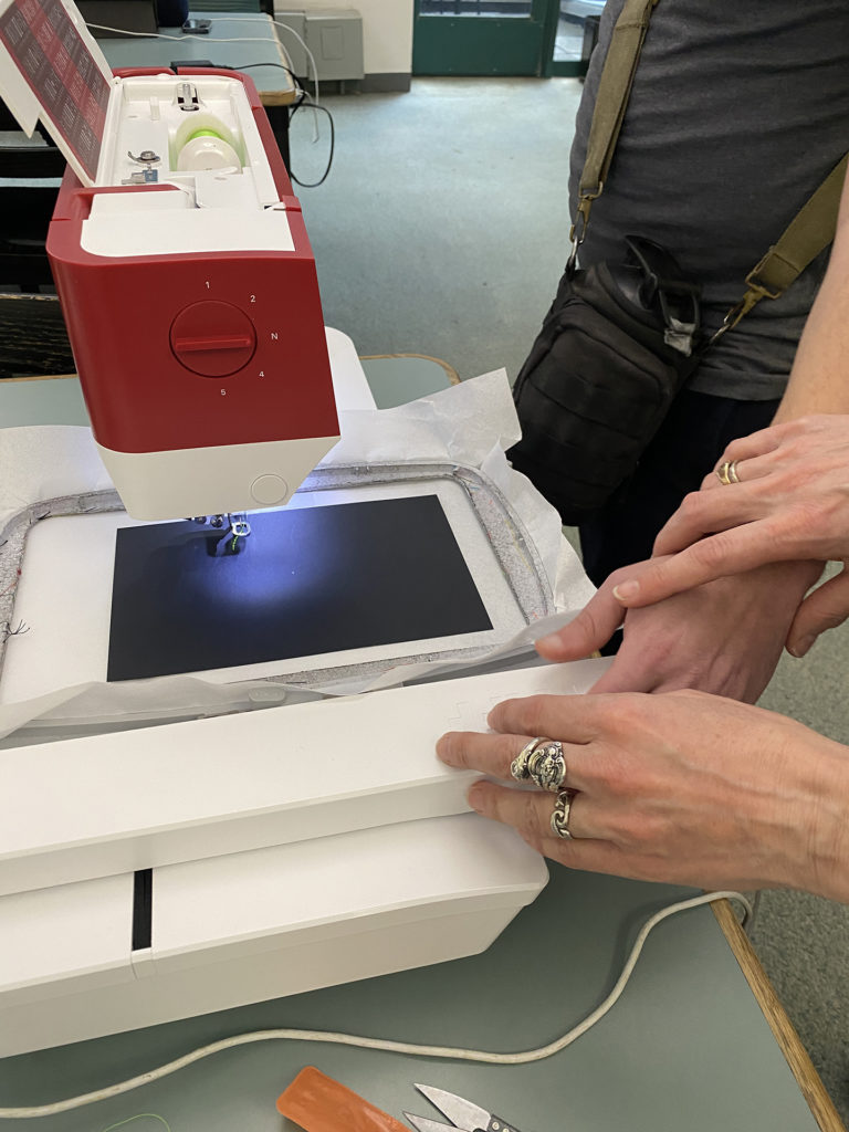 Two sets of hands guide a large embroidery hoop on to a digital embroidery machine. A black piece of paper is in the hoop