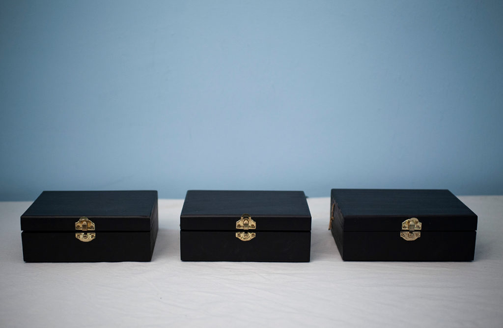 Photo of 'I Am Here You Are Safe', three small black boxes sitting closed on a white table in front of a pale blue wall