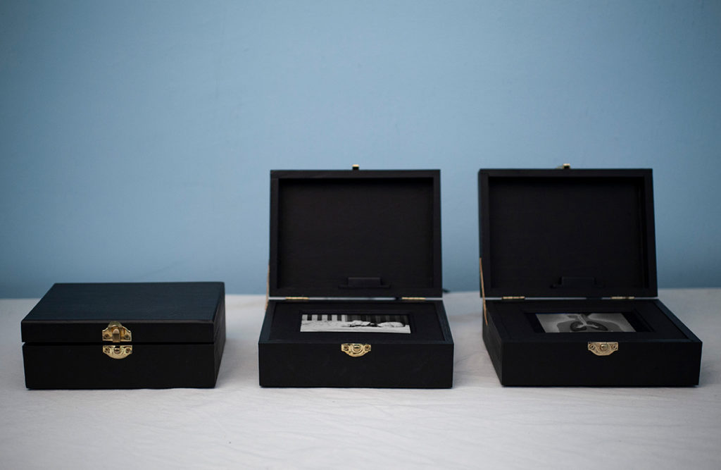 Photo of 'I Am Here You Are Safe', three small black boxes sitting with one on the left closed and the center and right one open on a white table in front of a pale blue wall