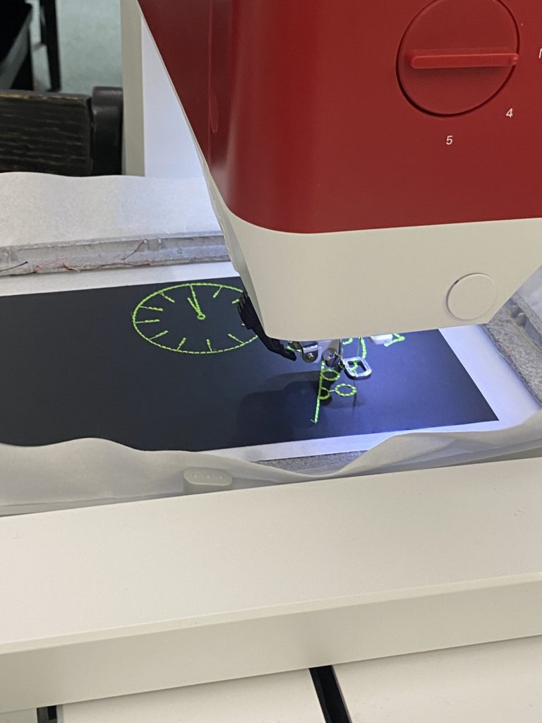 A digital embroidery machine stitches a clock image on black paper in bright green thread.