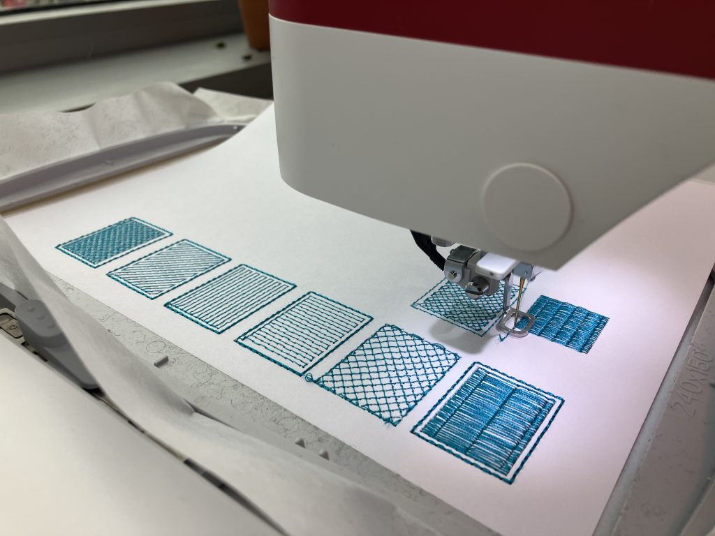 A close up of an embroidery machine stitching pattern swatches in blue thread on white paper.