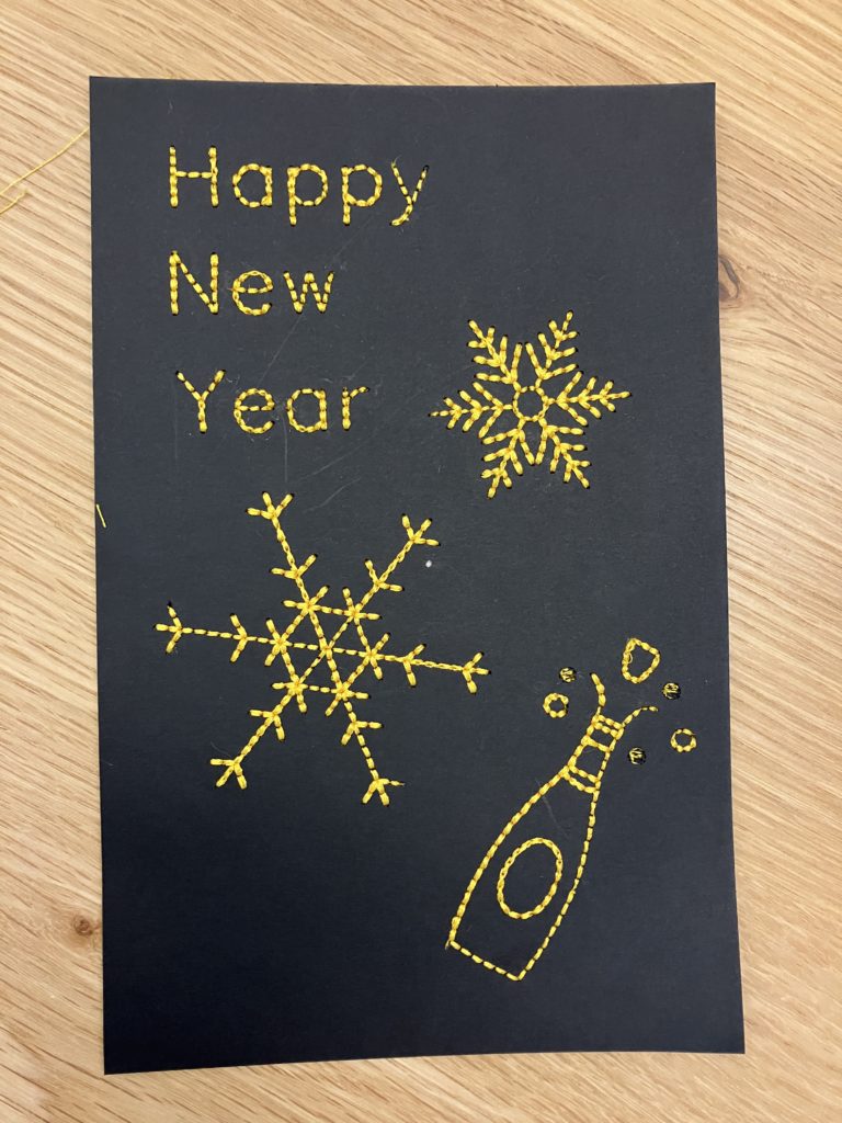 A Happy New Year's Card stitched in bright yellow thread on black cardstock. The text reads "Happy New Year". There is also small snowflake, a large snowflake, and a champagne bottle popping.