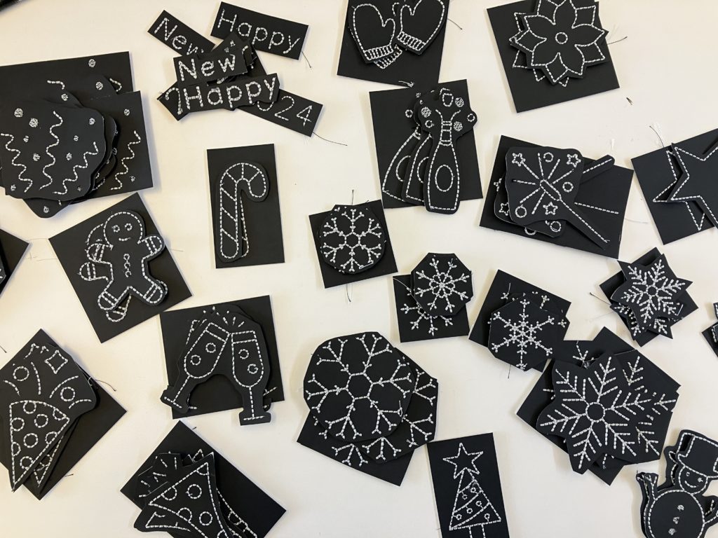 30 images including snowflakes, party horns, and the words Happy New Year stitched in silver thread on black card stock. The images are stitched on to squares of cardstock. 