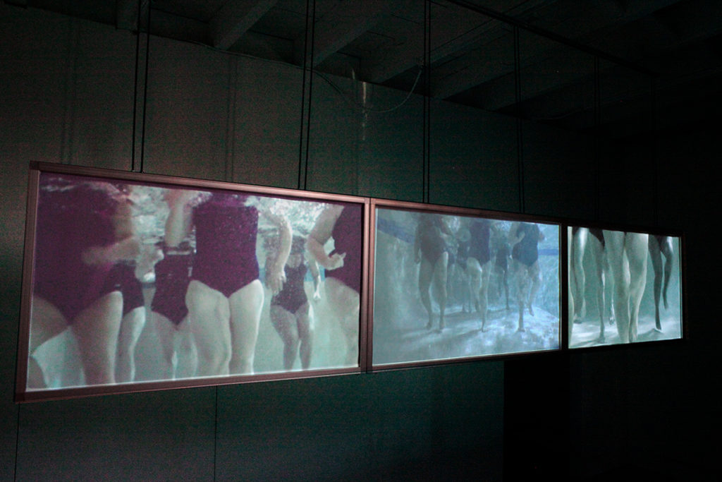 Photo of To The Deep (2013) A 3-channel underwater video - showing 3 screens hanging in a dark room with people swimming underwater