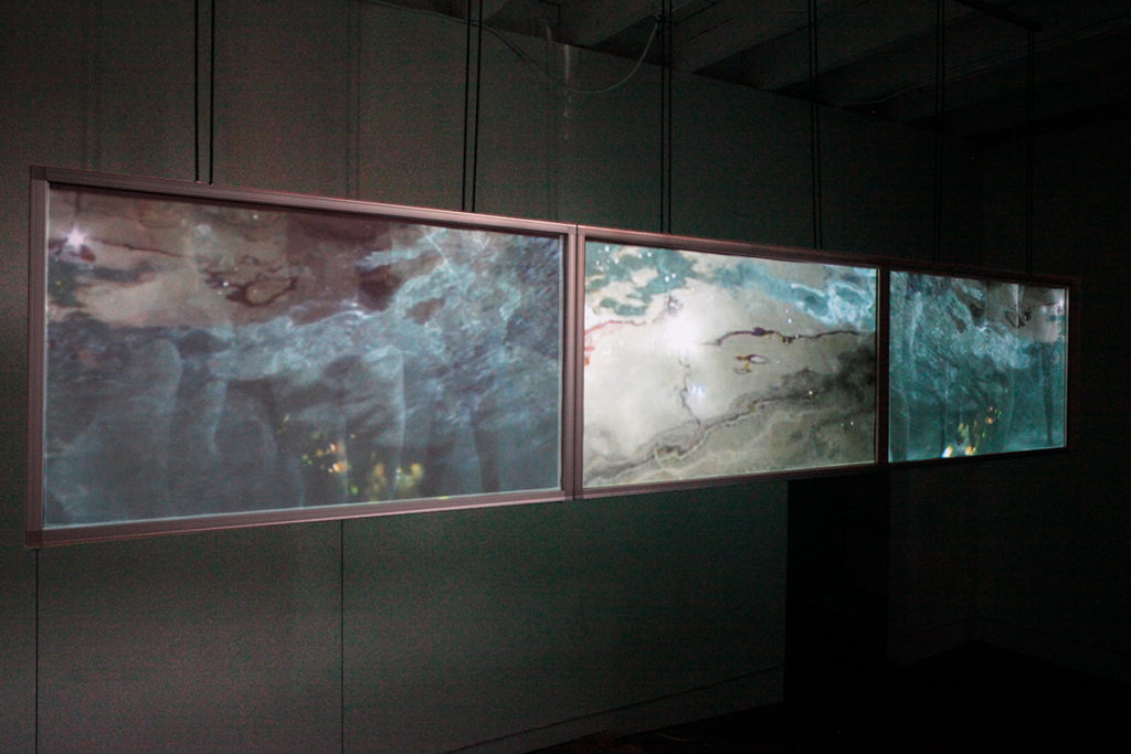 Photo of To The Deep (2013) A 3-channel underwater video - showing 3 screens hanging in a dark room with people swimming underwater