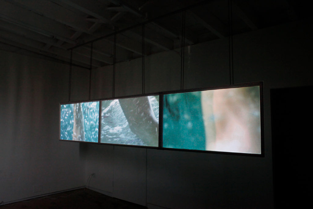 Photo of To The Deep (2013) A 3-channel underwater video - showing 3 screens hanging in a dark room with people swimming underwater